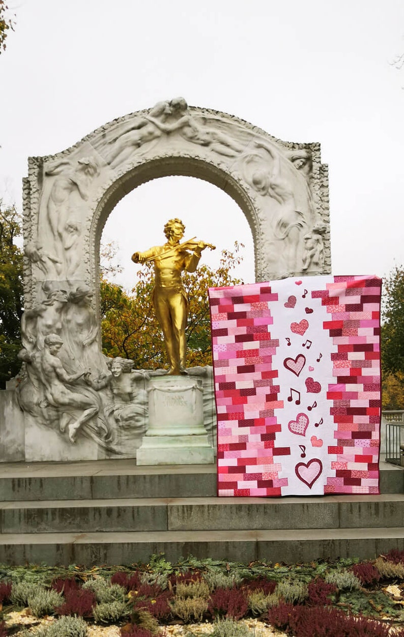 May include: A pink and red patchwork quilt with a white panel featuring hearts and musical notes. The quilt is displayed in front of a white stone archway with a golden statue of a man playing a violin.