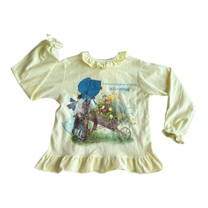 May include: A yellow long-sleeved shirt with a ruffled neckline and cuffs. The shirt features a vintage illustration of a girl pushing a wheelbarrow full of flowers. The text "A little love just grows and grows HOLLY HOBBIE" is printed on the shirt.