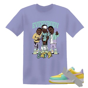 May include: A light purple t-shirt featuring a cartoon illustration of three people with the text "EVERYBODY" above them and "Eats B." below. A multi-colored sneaker is taped to the bottom right.