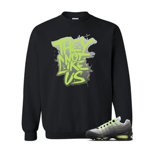 Puede incluir: Sudadera negra con el texto "THEY NOT LIKE US" en verde ne&oacute;n y gris. Un par de zapatillas deportivas grises, negras y verde ne&oacute;n est&aacute;n en la esquina inferior derecha.