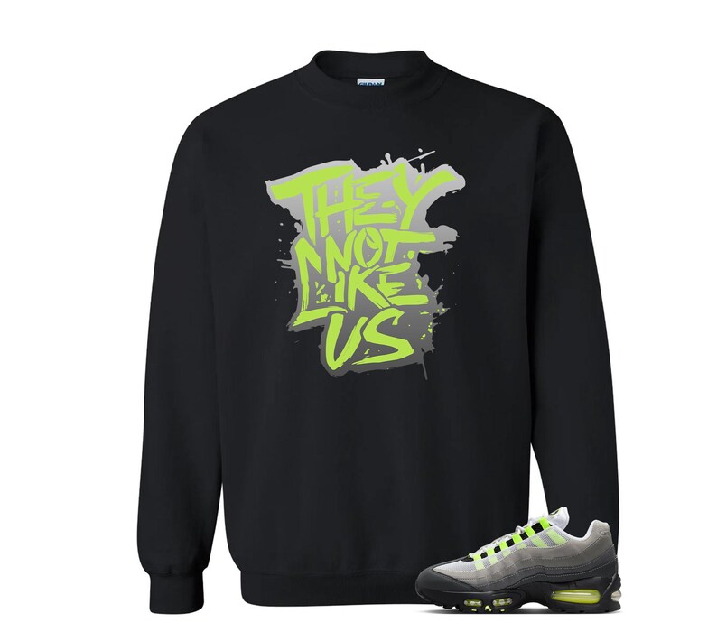 Puede incluir: Sudadera negra con el texto "THEY NOT LIKE US" en verde ne&oacute;n y gris. Un par de zapatillas deportivas grises, negras y verde ne&oacute;n est&aacute;n en la esquina inferior derecha.