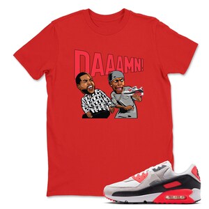 May include: Red t-shirt with the word "DAAAMN!" above a cartoon graphic of two men. One man is wearing a black and white checkered shirt, and the other is holding a sneaker. A gray, white, and red sneaker is in the foreground.