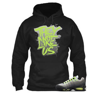 Puede incluir: Sudadera con capucha negra con el texto "THEY NOT LIKE US" en verde ne&oacute;n y gris. Un par de zapatillas deportivas grises, negras y verde ne&oacute;n est&aacute;n en primer plano. La sudadera tiene una capucha con cord&oacute;n y un bolsillo de canguro.
