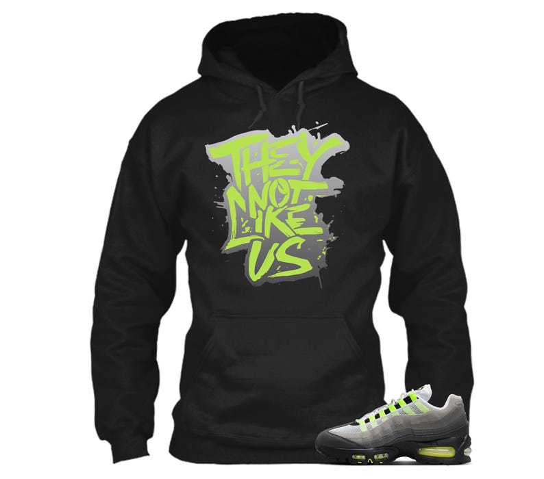 Puede incluir: Sudadera con capucha negra con el texto "THEY NOT LIKE US" en verde ne&oacute;n y gris. Un par de zapatillas deportivas grises, negras y verde ne&oacute;n est&aacute;n en primer plano. La sudadera tiene una capucha con cord&oacute;n y un bolsillo de canguro.
