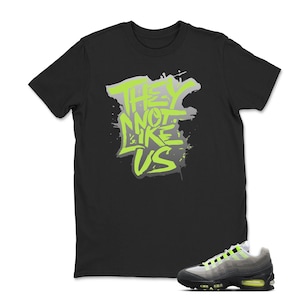 Puede incluir: Camiseta negra con el texto "THEY NOT LIKE US" en verde ne&oacute;n y gris. Un par de zapatillas grises y negras con detalles en verde ne&oacute;n en la parte inferior derecha.