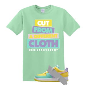 May include: A pale green t-shirt with the text "CUT FROM A DIFFERENT CLOTH" in stacked, colourful blocks. A multi-coloured trainer is taped to the shirt. The hashtag #BUILTDIFFERENT is below the text.