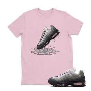 May include: A light pink t-shirt featuring a graphic of a sneaker with gray and black tones, appearing to splash into a puddle. A matching sneaker is placed below the shirt. The shirt is a crew neck.