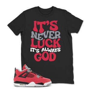 May include: Black t-shirt with the text "IT'S NEVER LUCK IT'S ALWAYS GOD" in red, gray, and white. A red and black sneaker with white and gray accents is in the foreground. The sneaker has a black sole and a red upper.