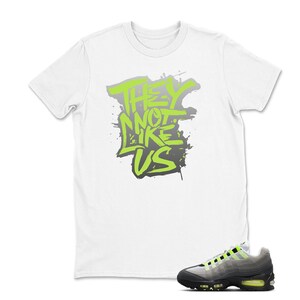 Puede incluir: Camiseta blanca con el texto "THEY NOT LIKE US" en verde ne&oacute;n y gris. Un par de zapatillas deportivas negras, grises y verde ne&oacute;n est&aacute;n en primer plano. Las zapatillas tienen una suela negra y detalles en verde ne&oacute;n.