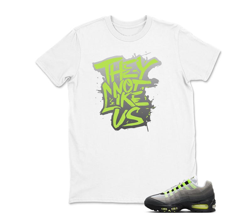 Puede incluir: Camiseta blanca con el texto "THEY NOT LIKE US" en verde ne&oacute;n y gris. Un par de zapatillas deportivas negras, grises y verde ne&oacute;n est&aacute;n en primer plano. Las zapatillas tienen una suela negra y detalles en verde ne&oacute;n.