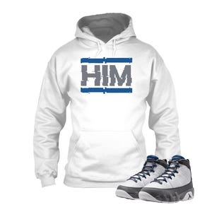 May include: White hoodie with the word "HIM" in gray and blue block letters. The hoodie has a front pocket and a hood. A pair of white, black, and blue sneakers are in front of the hoodie.