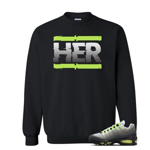 Pu&ograve; includere: Felpa nera con la scritta "HER" in un font in stile glitch, accentuata da linee orizzontali verde neon. Un paio di scarpe da ginnastica grigie, nere e verde neon sono in primo piano.
