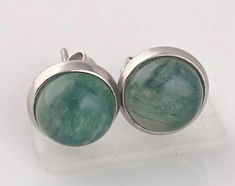 May include: A pair of round stud earrings. Each earring has a green stone set in a silver-coloured bezel. The stones have a marbled appearance with varying shades of green. The earrings are displayed on a white surface.
