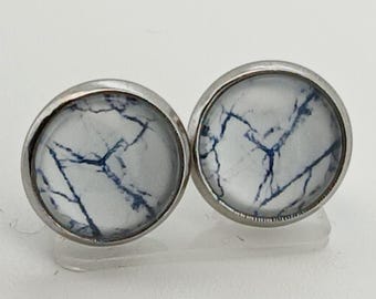 Könnte beinhalten: Ein Paar runde Ohrstecker. Jeder Ohrring hat einen weißen Hintergrund mit einem blauen Marmormuster, eingefasst in eine silberfarbene Metalleinfassung. Die Ohrringe werden auf einer weißen Oberfläche präsentiert.