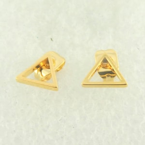 Puede incluir: Un par de pendientes de triángulo de color dorado. Los pendientes son huecos y tienen un diseño geométrico simple.