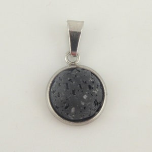 Op de afbeelding: Een ronde, donkergrijze lavasteen hanger met een zilverkleurige metalen rand en balein. De hanger heeft een textuur en wordt tegen een witte achtergrond getoond. Deze ketting is een sieraad.