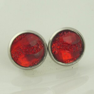 May include: A pair of round stud earrings with a bright red, textured center. The red stones are set in a silver-colored metal bezel. The earrings are displayed on a white surface.
