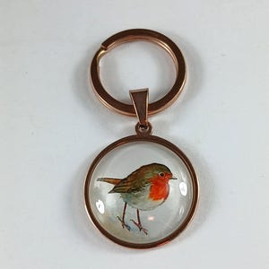 May include: A rose gold keychain with a round pendant featuring a detailed illustration of a robin. The bird has a red breast, brown wings, and is set against a clear background. The keychain is a charming accessory.