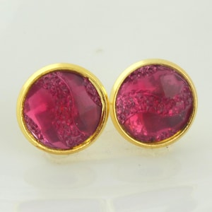 Op de afbeelding: Een paar ronde oorstekers met een goudkleurige zetting. Elke oorbel heeft een levendige, getextureerde, magenta-kleurige steen. De oorbellen worden gepresenteerd op een wit oppervlak, waarbij het ontwerp en de kleur van de sieraden worden getoond.