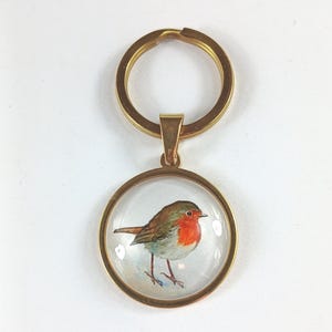 May include: A gold-colored keychain with a round pendant featuring a watercolor painting of a robin. The bird has a red breast, brown wings, and is set against a white background. The keychain is a simple, elegant accessory.