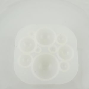May include: A white silicone mould with a square shape, featuring various sized circular cavities. The cavities are designed to create spherical or bubble-shaped objects. The mould is made of a flexible, translucent material.