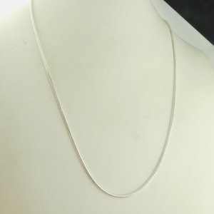 Pode incluir: Um delicado colar de prata exibido em um manequim branco. A corrente é fina e parece ser feita de pequenas ligações interconectadas. O colar é drapeado em uma curva suave, mostrando sua elegância simples.