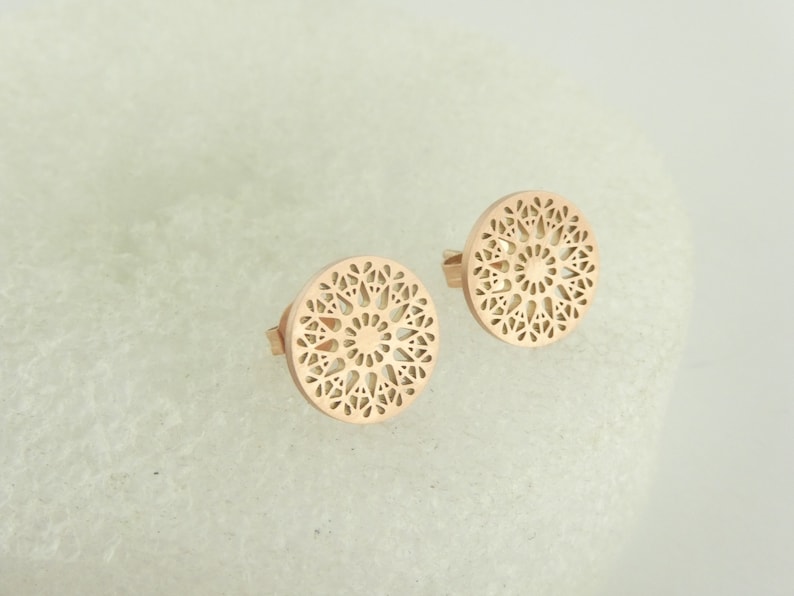 May include: A pair of rose gold earrings with a delicate, circular, mandala-like design. The earrings are made of metal and have a polished finish.