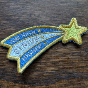 Puede incluir: Un parche bordado en forma de estrella fugaz, con un diseño azul y blanco y un contorno dorado. La estrella tiene un centro amarillo y el parche dice "Aim High Strive Higher".