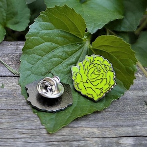 Puede incluir: Un pin de esmalte verde con una rosa estilizada y una parte posterior de pin de plata.