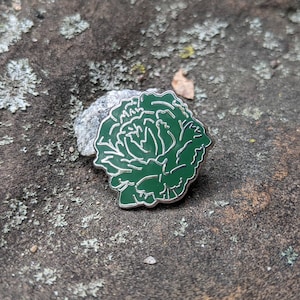 May include: A green enamel pin featuring a stylized floral design. The pin is shaped like a flower with multiple layers of petals.
