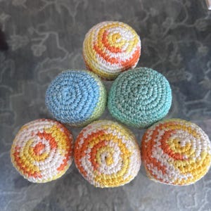 May include: A collection of seven crocheted juggling balls. The balls feature a spiral design in various colors, including yellow, orange, white, blue, and green. The balls are arranged in a pyramid shape.