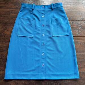 May include: A bright blue skirt with a button-down front and two front pockets. The skirt has a high waist and a straight silhouette. The fabric appears to be a smooth, woven material. The skirt is displayed on a wooden surface.