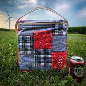 Puede incluir: Una bolsa de refrigeración de patchwork azul y rojo con un asa blanca. La bolsa de refrigeración está sentada sobre hierba verde frente a una lata de sidra de manzana Busch Light.