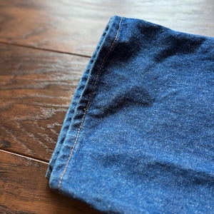 May include: Close-up of a section of blue denim fabric, folded over. The denim has a classic, slightly faded appearance. The edge is stitched with a contrasting orange thread, highlighting the seams. The fabric is resting on a wooden surface.