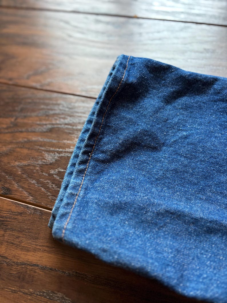 May include: Close-up of a section of blue denim fabric, folded over. The denim has a classic, slightly faded appearance. The edge is stitched with a contrasting orange thread, highlighting the seams. The fabric is resting on a wooden surface.