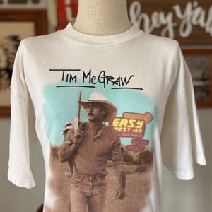 May include: Off-white t-shirt featuring a graphic of Tim McGraw in a cowboy hat, jeans, and a long-sleeved shirt. The text "Tim McGraw" is above the image. The background includes a sign that says "Easy Rest Inn".