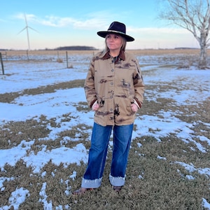 May include: A tan button-up jacket with a brown collar and a horse pattern. The jacket is paired with blue jeans and a black hat. The outfit is worn outdoors in a field with snow on the ground.