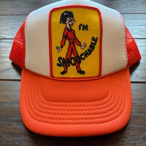 May include: Orange and white trucker hat featuring a cartoon graphic. The graphic shows a person in a red outfit with the words "I'M SMOOCHABLE" in black text on a yellow background. The hat has a curved peak.