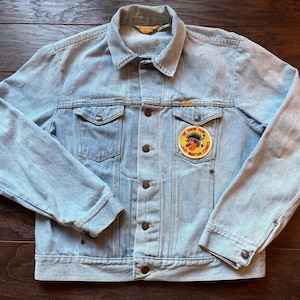May include: Light blue denim jacket with a button-up front, two chest pockets, and a collar. A circular patch on the chest reads "WE KNOW NOW JUST WHAT'M CHANCE" with a cartoon character. The jacket has a vintage look.