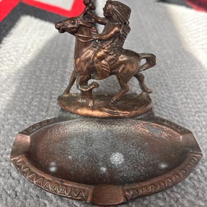 May include: A vintage bronze ashtray featuring a detailed sculpture of a Native American figure riding a horse. The ashtray has an oval shape with a decorative border and a central depression for ashes.