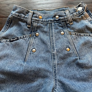 May include: A pair of light blue denim jeans with a high waist and a button fly. The jeans have a Wrangler logo on the back pocket and silver metal rivets along the seams.