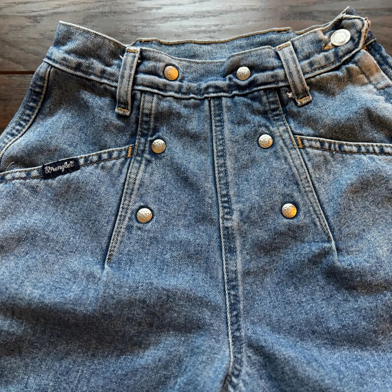 May include: A pair of light blue denim jeans with a high waist and a button fly. The jeans have a Wrangler logo on the back pocket and silver metal rivets along the seams.
