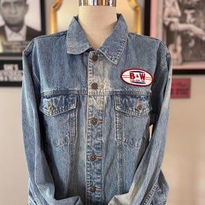 May include: A light blue acid-wash denim jacket featuring a classic collar, long sleeves, and a front button closure. An oval patch on the left chest displays the text B & W CO-OP, INC. in red and black lettering against a white background.