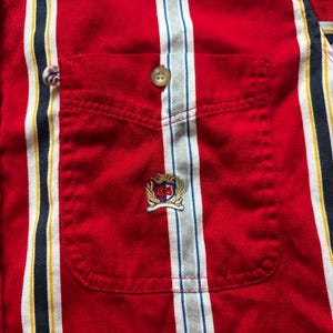 May include: Red striped button-down shirt with a chest pocket and a small embroidered logo. The logo is a shield with a crown and the letters TH.