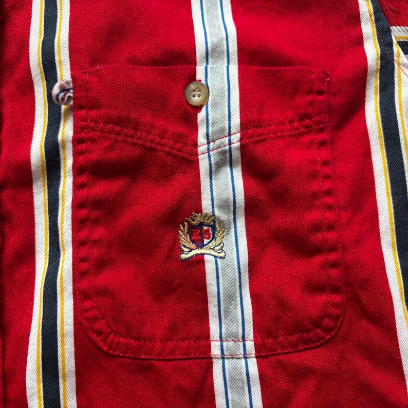 May include: Red striped button-down shirt with a chest pocket and a small embroidered logo. The logo is a shield with a crown and the letters TH.