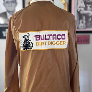 May include: A tan windbreaker jacket featuring a large rectangular patch on the back. The patch displays a black and white graphic of a motorcyclist and the purple and yellow text BULTACO DIRT DIGGER. The jacket has a white collar trim.