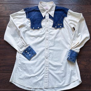 May include: Cream-colored long-sleeved shirt with denim accents. The shirt features denim shoulder yokes, cuffs, and a collar. Pearl snap buttons run down the front. The shirt has a Western style with embroidered initials on the yoke.