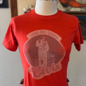 May include: A red t-shirt featuring a faded graphic of Elvis Presley with the words "Love Me Tender" and "Elvis". The shirt is displayed on a mannequin. Vintage apparel.