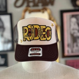 May include: A brown and cream trucker hat featuring a large yellow embroidered patch that reads RODEO with smaller text including GRATIOT CO. SHERIFF POSSE ITHACA MICH. Two small gold star accents are on the front panel above a dark brown brim.