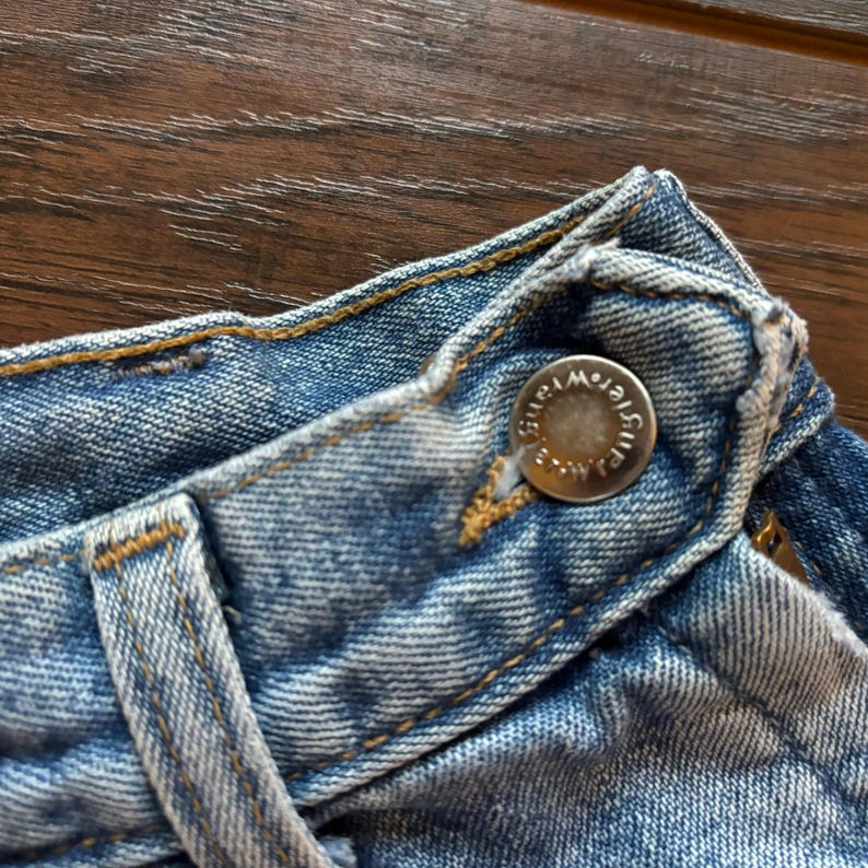 May include: Close-up of a light blue denim waistband with a silver button that reads "Guess" in a circular design.